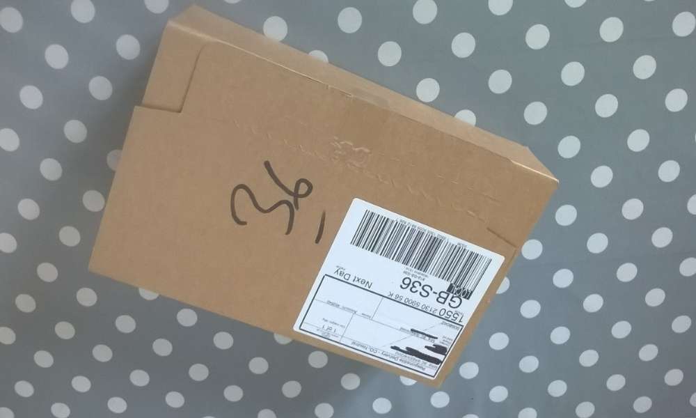 Image of a brown box with a white address sticker with the personal information crossed out in black marker pen, against a page grey and white polka dot background.