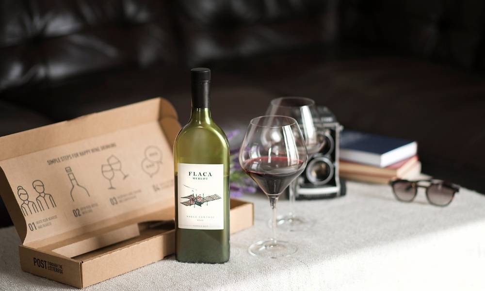 Image of a flat bottle of wine labelled Flaca Merlot in front of a brown box on a table covered in a white table cloth. To the right of the bottle are two wine glasses half full with red wine.